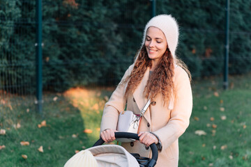 Young woman in a beige coat walks with a child in a stroller through the town. Mother with pram. Woman with baby stroller walks in the park at sunset.