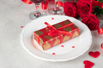 Gift in wrapping paper on white plate, red roses and two glasses for wine, concept for Valentines Day