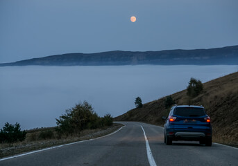 A blue car on a mountain road descends to the clouds in a mountain valley against the backdrop of a...