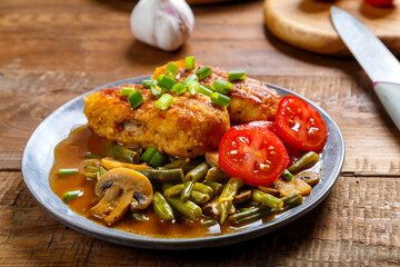 Chicken cutlets with green beans and mushrooms in tomato sauce in a plate next to garlic and a knife on the table.