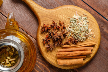 On the table on a wooden board anise, cinnamon, ginger and lemongrass grass next to a transparent tea pot top view.