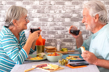 Cheerful caucasian senior couple toasting with red wineglass while sitting face to face at table having meal or brunch together at home