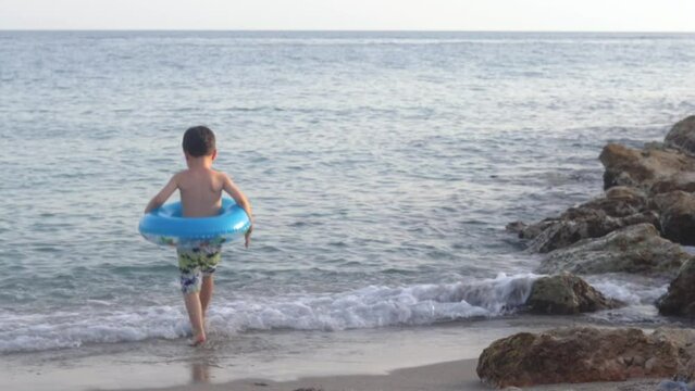 adorable kid boy preschooler playing having fun on seashore sand beach waves water ocean.child with blue inflatable ring around waist running in sea water,leaning on beach.palm print summer vacation