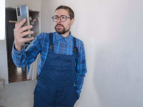 Young Man In Blue Work Suit Doing Repair Apartment. Home Renovation Concept. Man Having Video Call On Smartphone Over White Concrete Wall Background