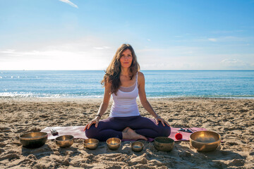 Female sound therapist with Tibetan singing bowls sitting in lotus pose, looking at the camera with...