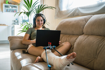 Young woman with broken leg lying on bed while using an laptop