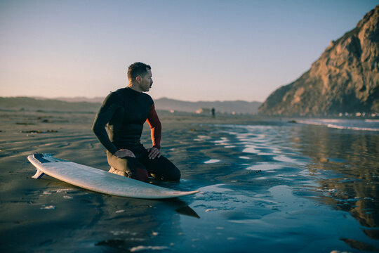 A surfer kneels on the beach next a surfboard. - Powered by Adobe