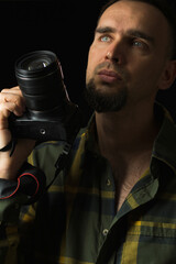 Portrait of a man with a camera. Black background. Photographer in green plaid shirt with beard holds camera at face level with lens up. Low key.