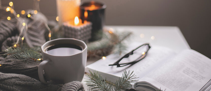 Banner image. Cozy home still life concept. Cup of hot coffee and opened book with atmospheric lights and burning candle. Winter holidays, Christmas time concept. Long banner for web site