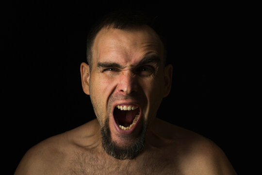 A bearded man screams emotionally with his mouth wide open. Portrait of a screaming man on a black background. Life problems.