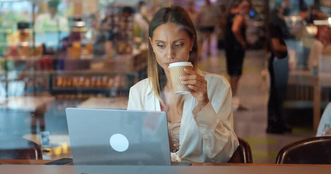 Confident young businesswoman, successful entrepreneur working on laptop remotely, sitting at wooden table in a cafeteria, drinking coffee while thinking on new project. Business. Telecommuting