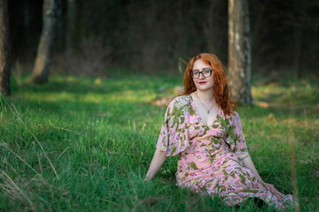 Portrait of a young beautiful red-haired girl in a dress sitting in the forest on the grass.