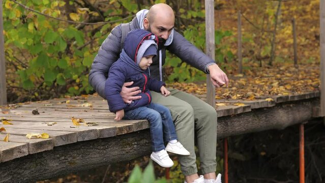 Bearded father and son are sitting on river bridge and looking at water flow. They talk, spend leisure time together. A man and 2 years old boy sit on a wooden bridge. Day vacation on river outdoors.