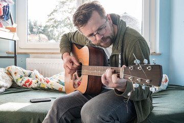 Man with an acoustic six-string guitar is learning a new tune while sitting on bed at home.