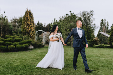 A stylish, young, fashionable groom in a blue suit and a beautiful, curly brunette bride in a white lace dress with a bouquet are walking, holding hands, in a park, garden. wedding photography.