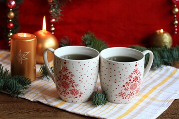 Christmas tea for two, mugs of herbal tea. Holiday decoration of golden candles,  coniferous tree branch and baubles on red background. Copy space.