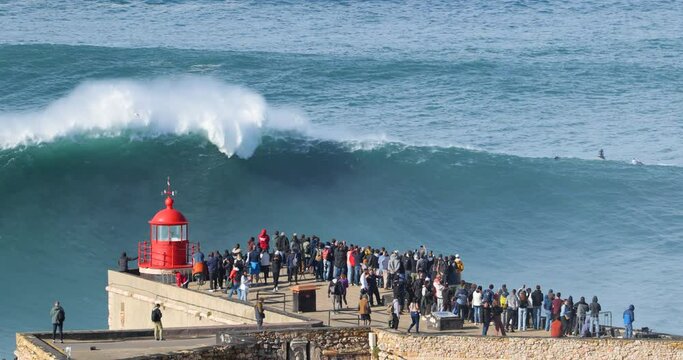 People watching the big giant waves crashing near the Fort of Nazare Lighthouse in Nazare, Portugal. Biggest waves in the world. Touristic destination for surfing and lovers of radical sports.
