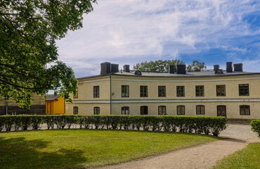 Helsinki, Finland - July 19, 2022: Suomenlinna Fortress. Yellow building at roundabout of Augustin Ehrensvärd's Grave under blue cloudscape. green lawn and foliage