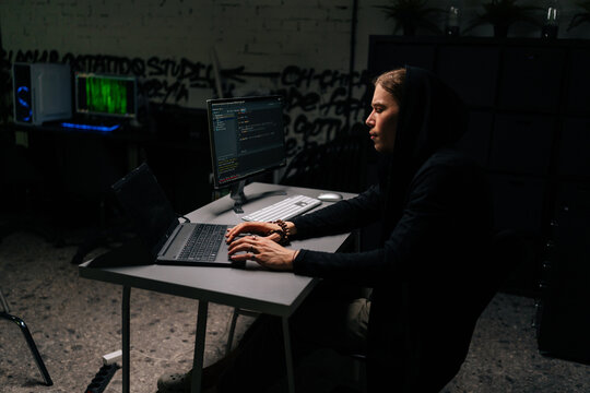 Side view of dangerous hacker man in sweatshirt hacking online web site, engaging hacking into security systems, using typing laptop and desktop computer. Concept of cyber attack, virus, malware