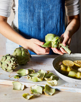 Person wearing blue apron peeling artichoke with bowl of leaves and lemons