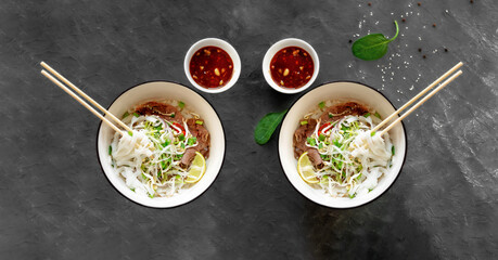 Pho bo, a traditional Vietnamese noodle and beef soup. Two cups of a delicious and hot dish with rice noodles on a dark gray background. Eating natural healthy Vietnamese food