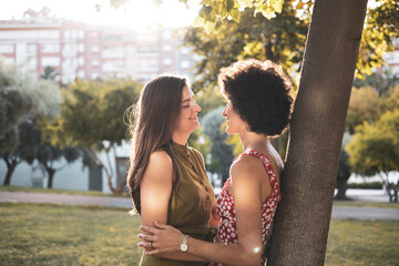 A young lesbian couple in a park. The girls look at each other before kissing by a tree, they spend time together, LGTB concept, lifestyle.