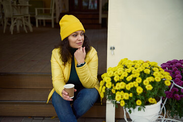 Pensive millennial multi-ethnic woman in bright yellow hat and jacket, sitting on the wooden...