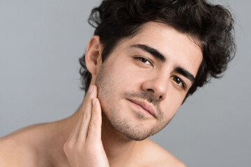 A young caucasian man with stubble beard is moisturizing face and skin with cosmetic oil while...