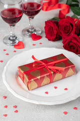 Gift in wrapping paper on white plate, red roses and two glasses wine on light table, concept for Valentines Day