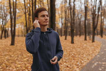 Athletic male runner listening to music in wireless earphones during training in autumn park 