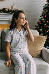 Preteen child in pajama talking on smartphone on couch during Christmas celebration at home