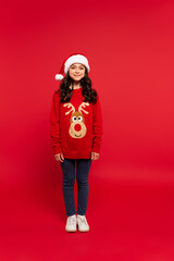 full length of happy girl in Christmas sweater and santa hat standing on red