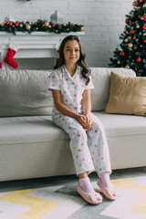 Smiling girl in dotted pajama sitting on couch near blurred fireplace and Christmas tree at home