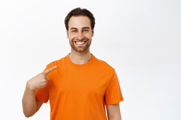 Portrait of young enthusiastic man smiling, pointing at himself, wearing orange t-shirt, white...