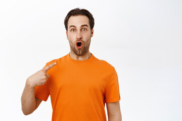 Fototapeta premium Portrait of young enthusiastic man looking surprised, pointing at himself, wearing orange t-shirt, white studio background