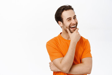 Image of charming brunette man with beard, winking and smiling at camera with pleased face, standing over white background