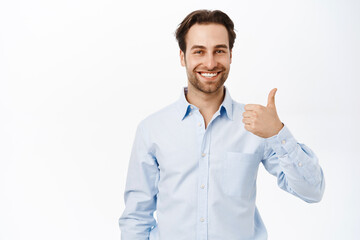 Handsome european businessman shows thumbs up, recommends smth good, approve and like brand or...