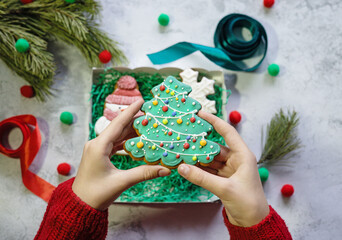 Girl's hands in red knitted sweater put Christmas tree in the form of snowman into gift box with gingerbread and snowflake gingerbreads next to pine branch, ribbons and wrapping paper. Christmas and N