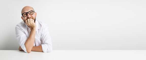 Smiling young man holding hand under chin while sitting at table on white background. Banner