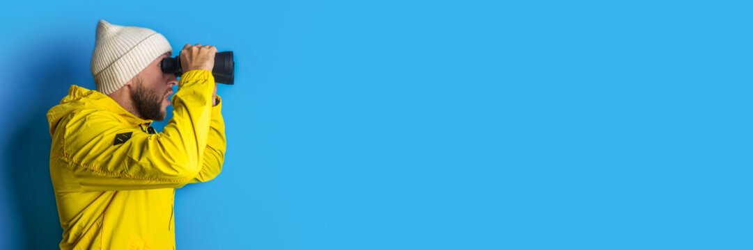 young man in a yellow jacket looks through binoculars on a blue background. Banner - Powered by Adobe