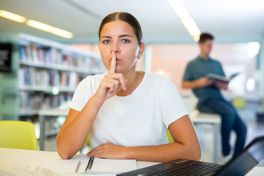 Serious young female student placing fingers on lips with shhh while using laptop in the library. High quality photo