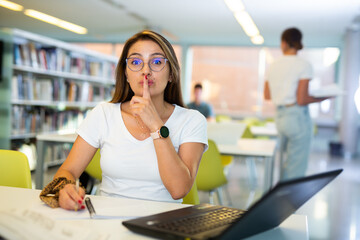 Young woman with glasses calls for silence in the public library - puts his index finger to his lips