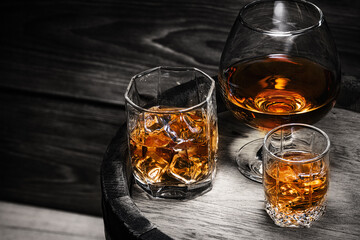 Cognac, whiskey, brandy poured into glasses stand on wooden barrel with alcoholic beverage whiskey close-up. Alcoholic beverages in glasses with ice on black background.