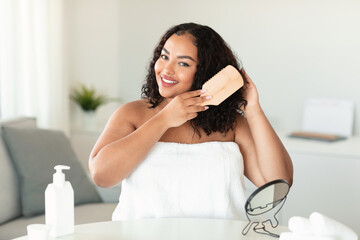 Pretty black chubby woman combing her curly hair with bamboo brush at home, sitting in bedroom and smiling at camera
