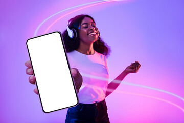 Positive black woman showing cell phone, using wireless headphones, mockup
