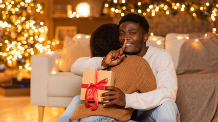 Happy young black wife hug husband with gift box, man show shh sign in cozy living room interior