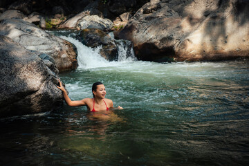 Fototapeta na wymiar Happy beautiful woman swimming in tropical mountain river with cascade waterfall. Young female traveller bathing in icy water for health improvement and strengthening of immunity.