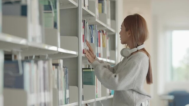 Asian Female student taking notes from a book at library. Young asian woman sitting at table doing assignments in college library.