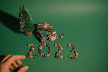 Green Christmas background with decorative pine tree, gifts boxes and female hand. Merry Christmas and Happy New Year Greeting Card with word 2023 made of colorful candies. Winter holidays wallpaper.