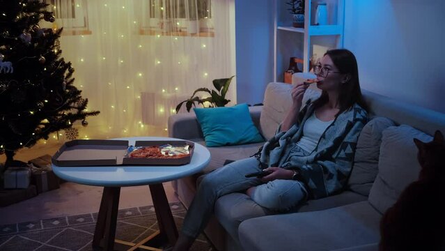 Young woman and pet cat eating pizza alone while watching tv, switch TV channels on the remote control at christmas on christmas tree background